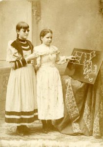 Photo of Edie Thomas a deaf-blind student and her blind friend standing before a propped up map of Massachusetts. Edie points to Hampshire County, 1895. Courtesy, Perkins School for Blind Archives