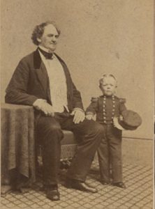 Photo portrait of George Washington Morrison Nutt standing with his hand on P.T Barnum’s knee, 1862. Barnum is sitting, Nutt is not quite as tall as the bend in Barnum’s elbow. Courtesy Barnum Museum