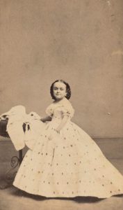 Photo portrait of Lavinia Bump, 1863, wearing short sleeved patterned dress and styled hair, side view near table, but Lavinia faces camera. Courtesy, Barnum Museum