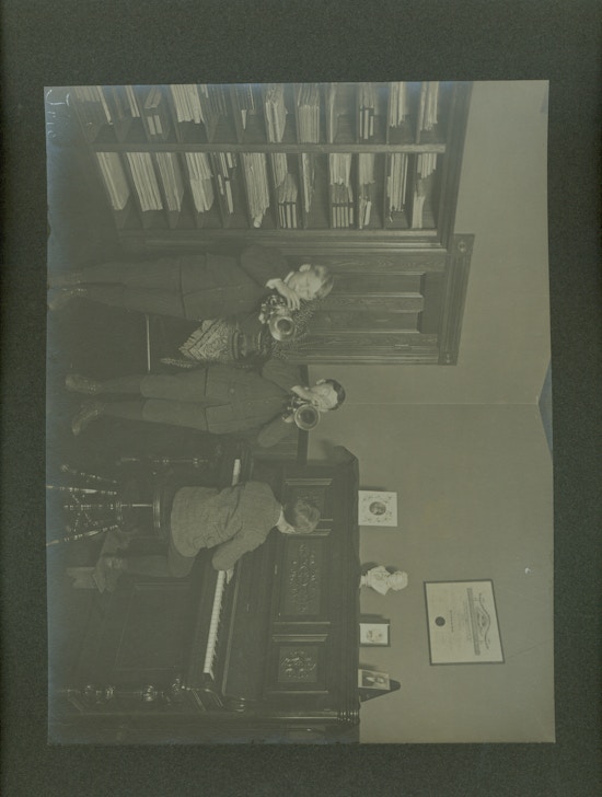 Two boys in dark suits playing trumpets, one boy in dark suit playing piano.