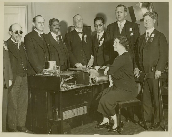 Keller sitting with hands on Visagraph, braille machine, men in suits stand around machine, Robert Irwin far right