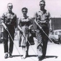 Three blind people walk down a city street, two men flanking a woman. Each carries a white cane in their right hand.