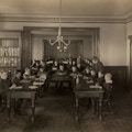 Classroom filled with boys at the Perkins Institution and Massachusetts School for the Blind in South Boston. They are seated around a large U-shaped wooden table with an assortment of plants, pine cones, and flowers. Each boy has a writing tablet and writing instrument.
