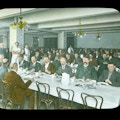 Large dining hall, men serving food to men with suit jackets at long tables, tablecloths set.