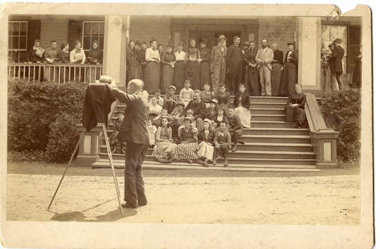 Behind the scene photograph of the group photograph of the first summer American Association To Promote Teaching Speech To The Deaf meeting taken on a Lake George porch with Alexander G. Bell. Photographer and camera are positioned left with the meeting participants in the background.