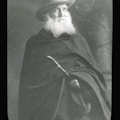 Alexander Graham Bell, facing right, wearing dark cloak and hat, grayed beard, holding cane or stick.