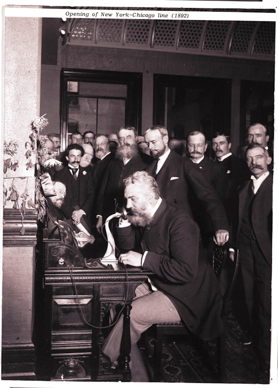 Alexander Graham Bell speaks into a telephone.  A group of men in suits look on.