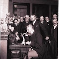 Alexander Graham Bell speaks into a telephone.  A group of men in suits look on.