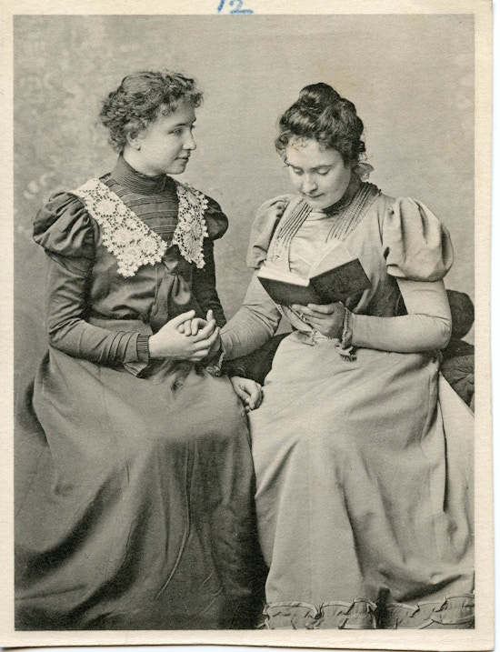 Anne Sullivan (right) finger spelling a book with Helen Keller (left), both seated.