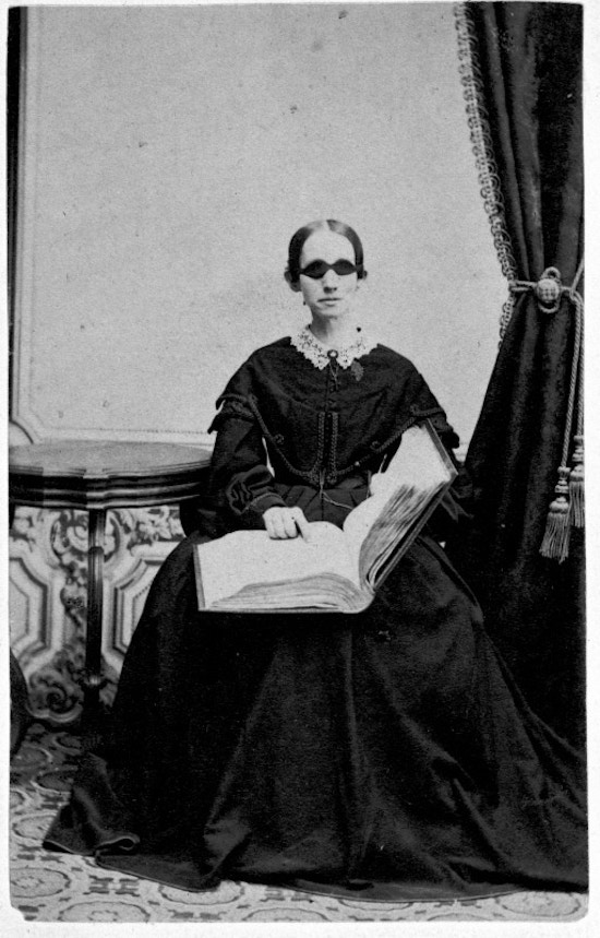 Bridgman, seated facing camera, holding book, dark dress with lace collar, dark glasses.