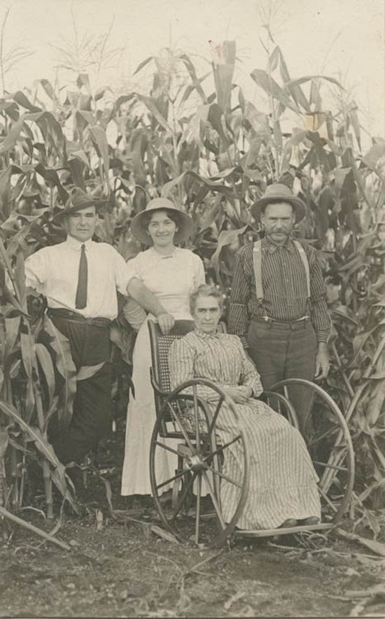 Two men and one woman standing behind elderly woman in a wheelchair, tall corn in background.