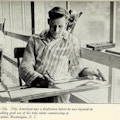 Young man sits at drafting table next to large window.