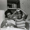 A boy at a kitchen table holds his hands on top of his head and smiles while a woman looks on.