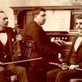 Portrait of three blind musicians seated with their instruments -- a violin, a clarinet, and a piano.