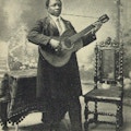 A black man wearing long coat and necktie stands playing a guitar in front of a table and a chair