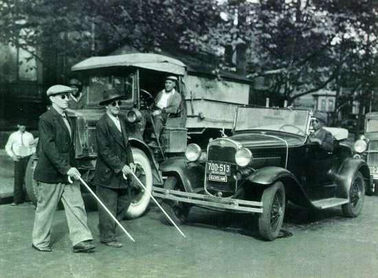 Photograph of two blind men, using white canes, crossing the street in front of a truck and a car.