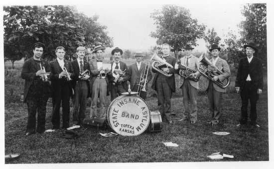 A nine-piece brass band stands in a field in front of trees holding their instruments.  They stand around a large drum on which is printed "State Insane Asylum Band, Topeka, KS".