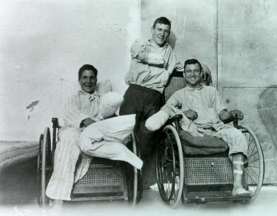 Three smiling veterans. Two leg amputees are seated in wicker wheelchairs. An arm amputee stands behind them.