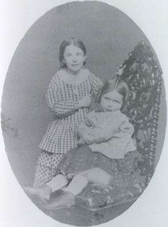 Photograph of two young girls, one seated in a chair and the other standing behind her.