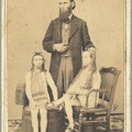 An average-sized man stands between two sitting short-stautured men with long hair and beards.