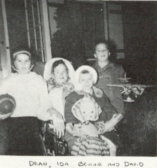 Ida Brinkman, in a wheelchair, surrounded by a boy holding a football, a girl holding a doll, and a boy holding an airplane.