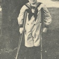 A young boy dressed in a sailor's suits stands with crutches and smiles.