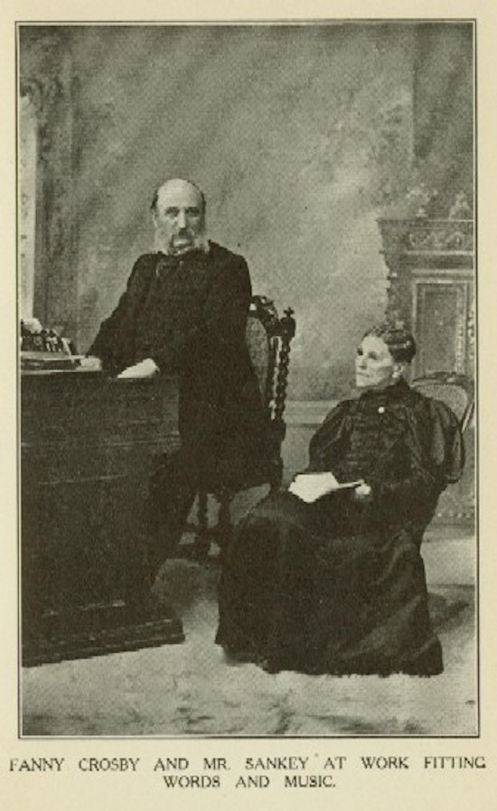A man sits at a organ, and a woman in dark glasses holds a book.