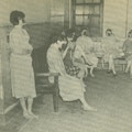 Seven women, most barefoot, sit on benches in a room.