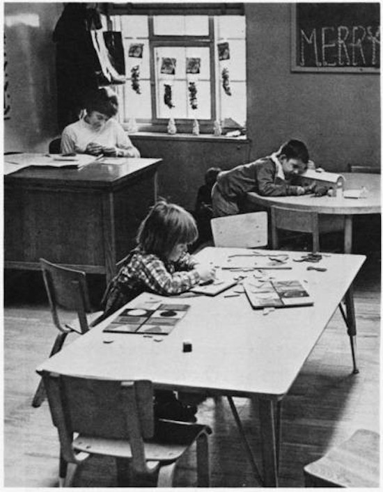 A teacher at her desk and two children at tables.