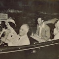 Franklin Roosevelt drives a car as King George speaks to him from the back seat.  Roosevelt is seated next Queen Elizabeth.