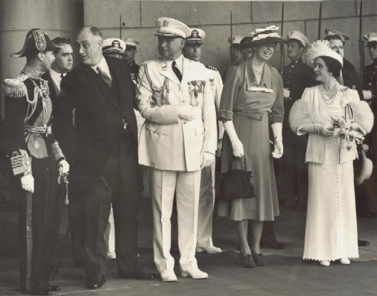 Roosevelt, holding the arm of a man in uniform, speaks with the King.  Eleanor Roosevelt and Queen Elizabeth stand nearby.