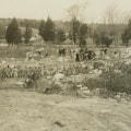 A row of men, with horses and wagons in background, work clearing the ground of debris.