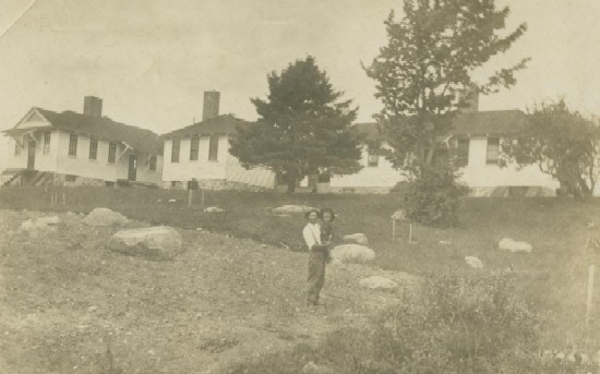A man holds a boy in a rocky field before several small white buildings.
