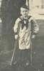 A young boy dressed in a sailor's suits stands with crutches and smiles.