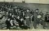 A group of civilian men sit on benches as two men in military uniforms hand out exams