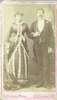 A portrait of a couple standing, the woman in a dress with ruffles and the man in a suit.
