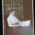Keller seated in chair in front of bookshelf reading a raised-print book.