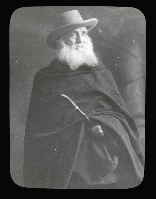 Alexander Graham Bell, facing right, wearing dark cloak and hat, grayed beard, holding cane or stick.