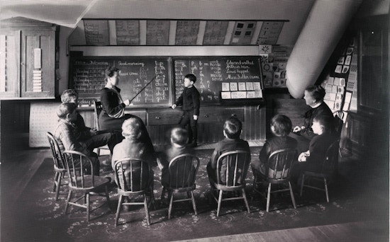 Photograph of Sarah Fuller's class with seated students facing chalkboard, away from camera. One student is standing at the board with Fuller instructing.