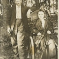 Woman sitting in wheelchair and holding crutch. Man stands next her holding cigar.