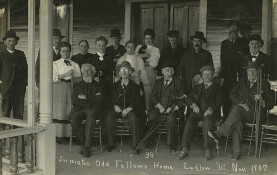 Group of elderly men, some with canes and crutches, sitting and standing on porch.  Also five women.