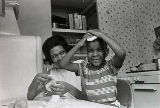 A boy at a kitchen table holds his hands on top of his head and smiles while a woman looks on.