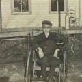 Photograph of a boy using a wheelchair in the yard of a small house.