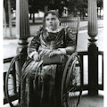 A young girl, holding a book, sits in a wheelchair on a porch.