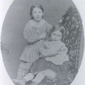 Photograph of two young girls, one seated in a chair and the other standing behind her.