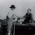 President Roosevelt sits up on the back of a car seat and shakes hands with a farmer in overalls standing next to the car.
