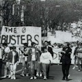 A crowd of protesters marches down a street.  A man holds a banner that reads The Board Busters.