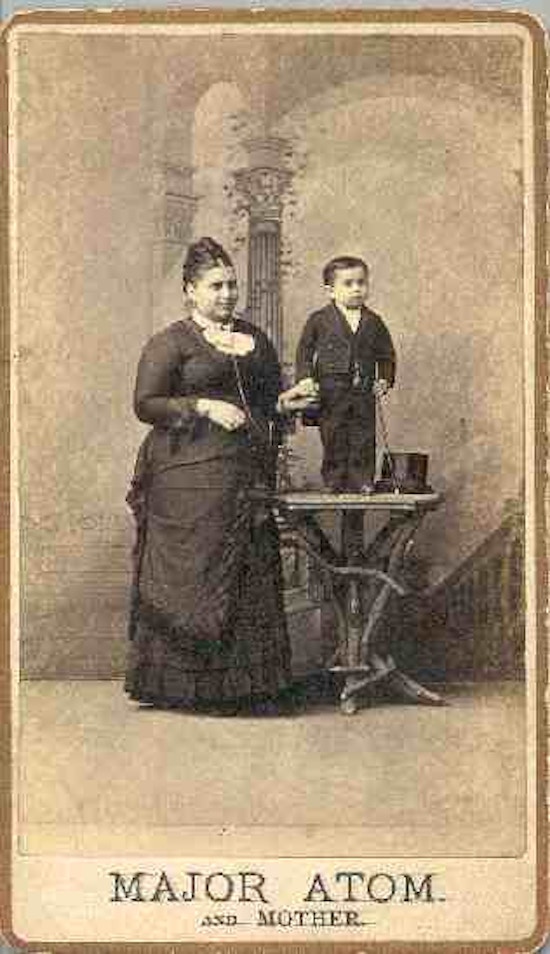 Major Atom, holding a top hat, stands on a table next to an average sized woman.