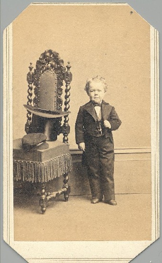 A short-statured young man in formal attire stands next to a chair.
