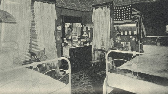 A bedroom with two beds, curtains, wallpaper, carpet, and an American flag.